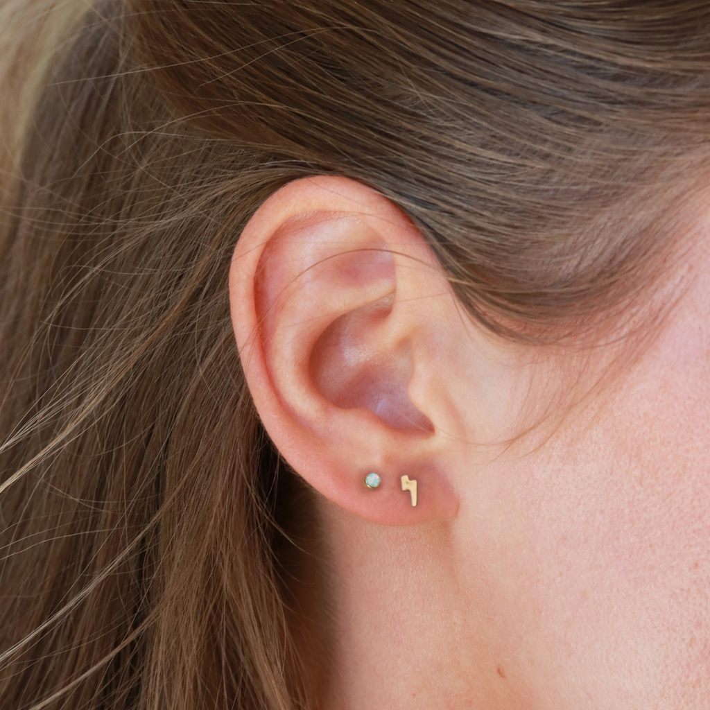A close-up view of a person's ear with two small gold earrings, one shaped like a lightning bolt and the other a heart-shaped charm.