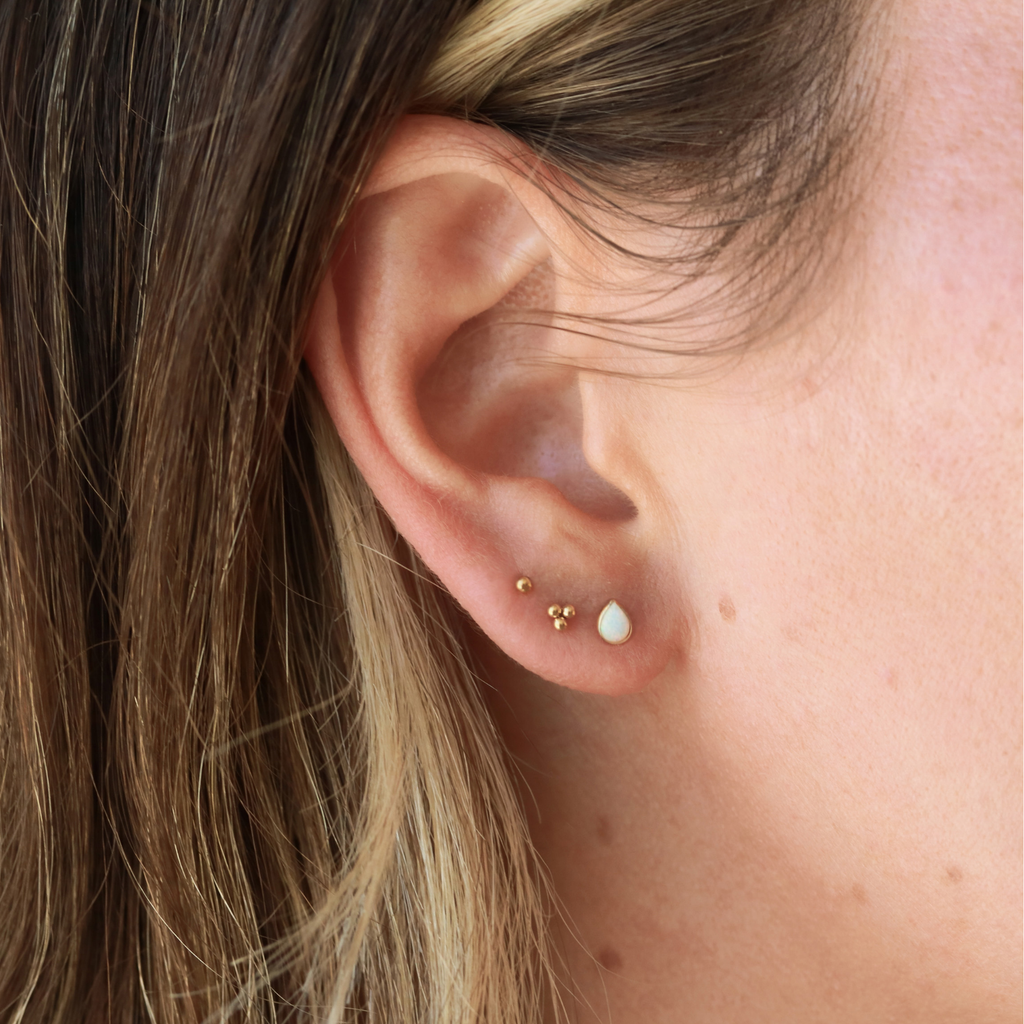 A close-up view of a person's ear adorned with two small gold earrings, one with a small white stone and the other with a small gold bead.